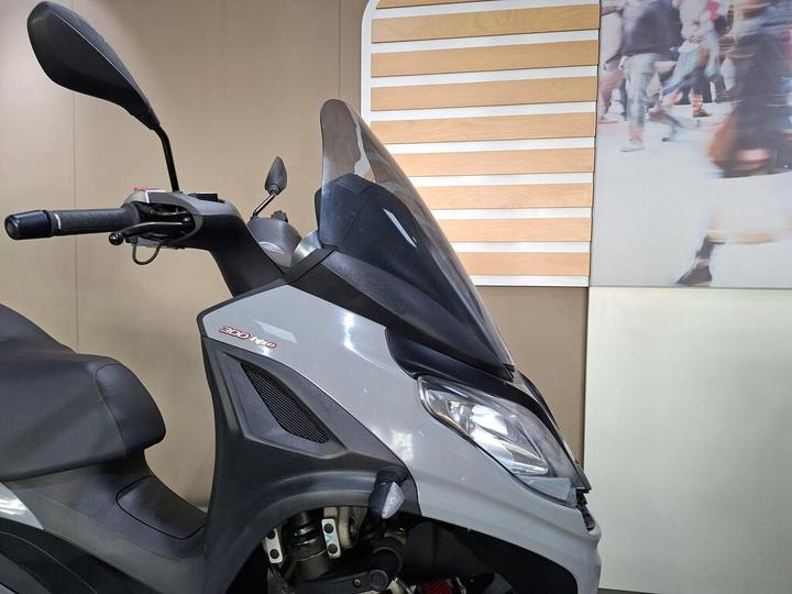 A close-up view of the handlebars and front of a gray scooter, featuring a transparent windscreen, control levers, and a partially visible seat. In the background, a blurred image of pedestrians is visible.