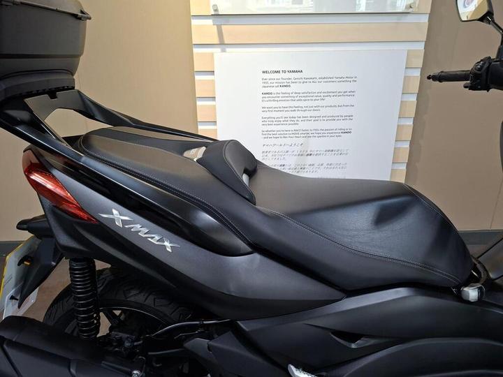 A close-up view of a Yamaha X-Max scooter's seat and rear section, showcasing its sleek black design and logo, with a simple backdrop featuring a welcome message about Yamaha.