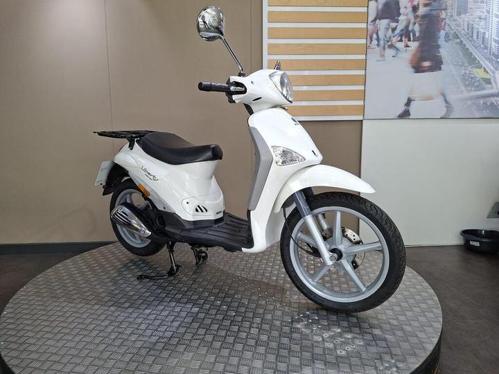 A white scooter displayed on a circular, metallic platform against a neutral background, featuring a sleek design with a single headlight and a comfortable seat.