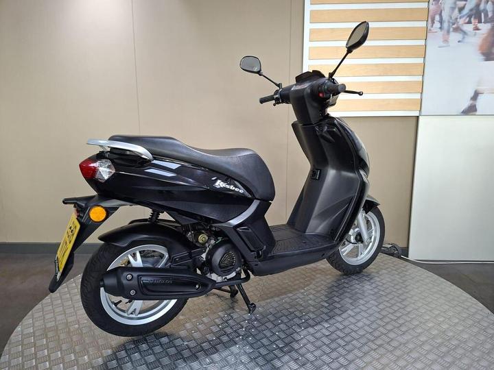 A black scooter parked on a textured platform, showcasing the side view with a focus on its sleek design and features. The background displays a neutral-toned wall with a glimpse of a patterned surface.