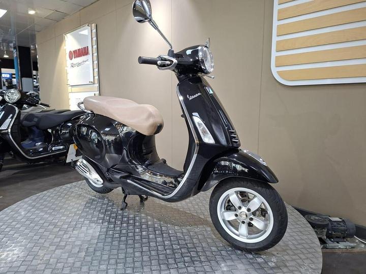 A black Vespa scooter with a brown seat is displayed on a circular platform in a showroom, with another motorcycle visible in the background and signage on the wall.