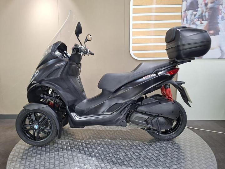 A sleek black three-wheeled scooter with a transparent windscreen, featuring stylish design elements and a rear storage compartment, on a textured platform against a neutral backdrop.