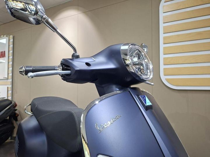 Close-up view of a matte blue Vespa scooter's front, showcasing the handlebar, mirror, and headlight, with a wooden panel background.