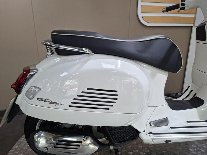 A side view of a white scooter featuring a rounded body, a black seat with silver trim, and a chrome rear grab rail. The scooter displays vent-like designs on the side and has a polished exhaust. The background features a simple beige wall.