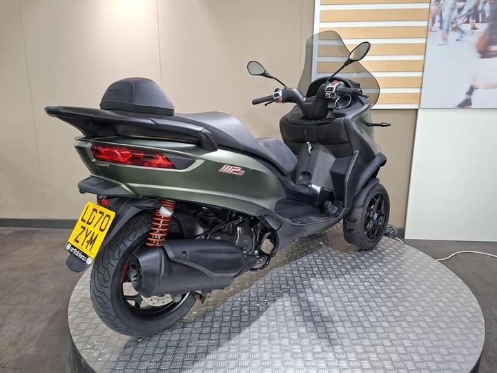 A three-wheeled scooter with a sleek, modern design parked on a circular platform, featuring a green body, dual rear lights, and a black seat.