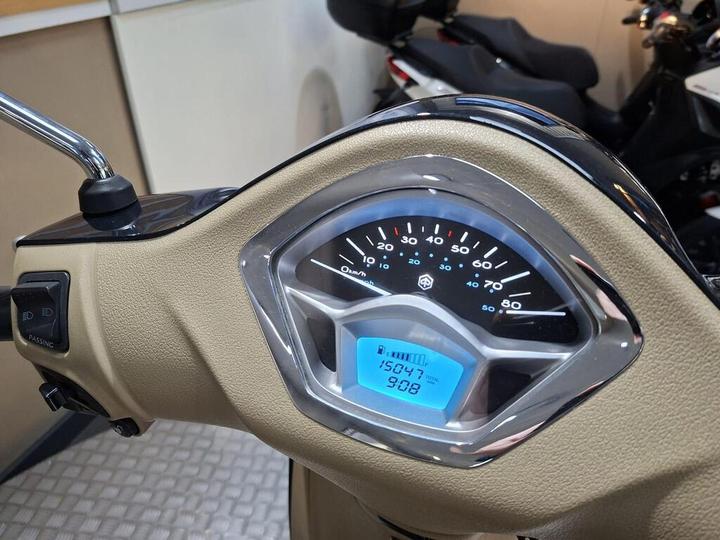 A close-up view of a scooter's dashboard displaying a speedometer and odometer. The speedometer has a black face with white markings, and the odometer shows a reading of 15047 km with a digital display below it indicating 908. The dashboard has a tan casing and chrome accents.