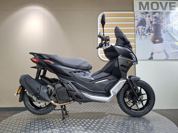 Aprilia SR GT 125 (2024) A black and silver scooter displayed on a textured circular platform, featuring a sleek design with a high front shield, a comfortable seat, and modern handlebars. The background includes a light-colored wall with a "MOVE" sign, and a blurred image of people walking.