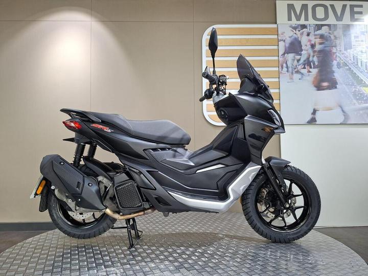 Aprilia SR GT 125 (2024) A black and silver scooter displayed on a textured circular platform, featuring a sleek design with a high front shield, a comfortable seat, and modern handlebars. The background includes a light-colored wall with a "MOVE" sign, and a blurred image of people walking.