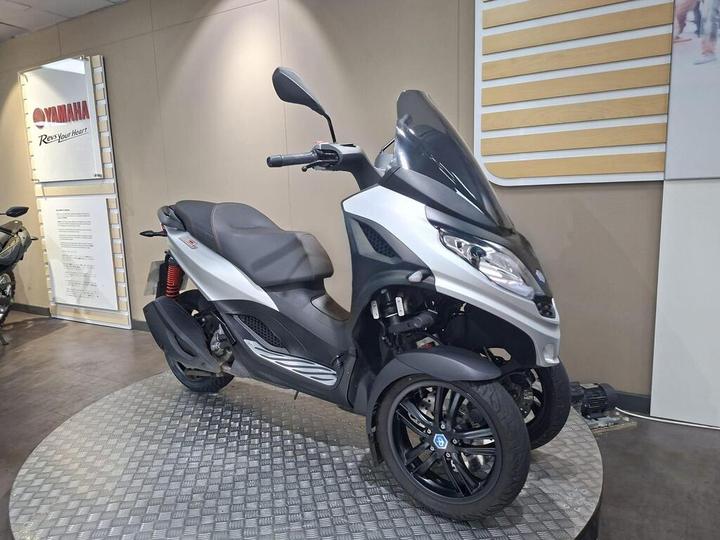 A silver three-wheeled scooter displayed on a circular platform in a showroom, with a modern design featuring a windscreen and black wheels. The background includes a Yamaha sign and promotional material.