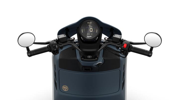 A top view of a scooter's handlebar, featuring a digital dashboard display at the center, with controls on both sides, including mirrors, throttle, and various buttons. The color scheme is primarily dark blue with a textured surface.