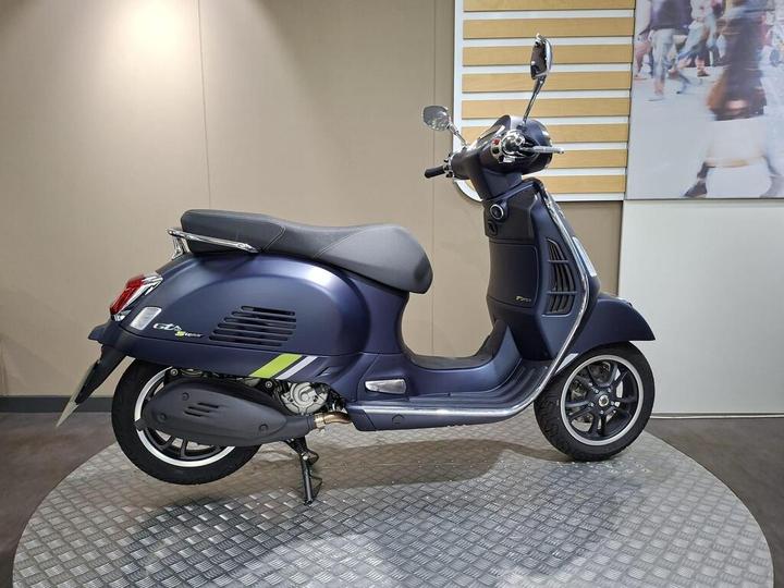 A blue and green scooter stands on a circular metal platform against a neutral backdrop, showcasing its sleek design and chrome details.