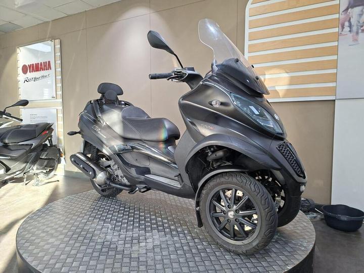 A black three-wheeled scooter displayed on a circular platform in a showroom, with a Yamaha logo visible in the background. The scooter features a windshield, sleek design, and modern styling.