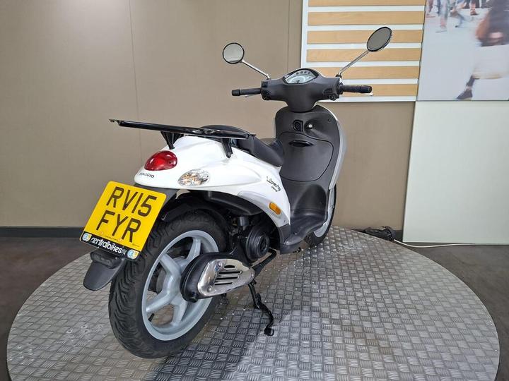 A white Piaggio scooter is displayed on a circular stand, featuring a rear view with a visible license plate reading "RV15 FYR." The background includes a neutral-colored wall and a partial view of an abstract image.