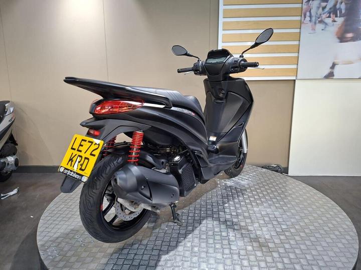 A black scooter with red shock absorbers is displayed on a rotating platform against a neutral background, showcasing its sleek design and features. The license plate reads LE72 KPD.