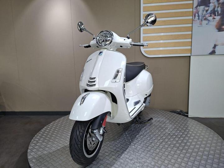 A white scooter standing on a circular platform in a showroom, featuring a sleek design with a round headlight and chrome accents. The background is a neutral beige with a hint of a patterned wall.