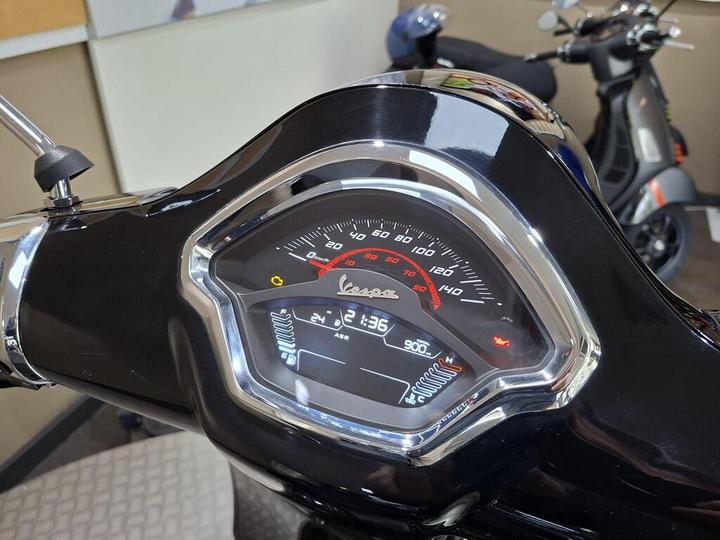 Close-up of a Vespa scooter's dashboard, featuring a speedometer and various indicator lights, with another scooter visible in the background.