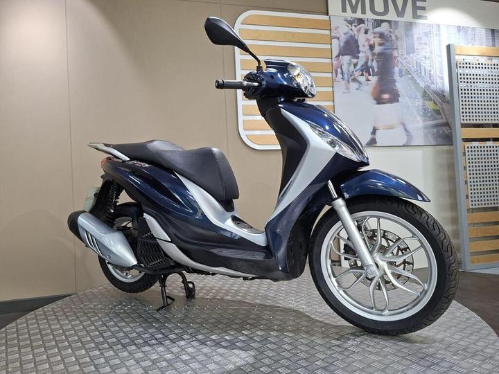 A blue and silver scooter is displayed on a circular platform against a neutral background, with an urban-themed backdrop featuring blurred pedestrians.