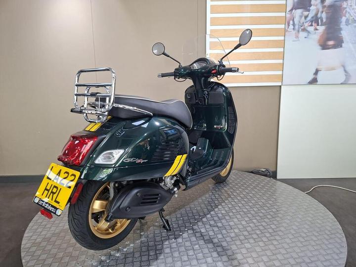 A green scooter with gold accents is displayed on a circular platform, featuring a luggage rack and a partial view of a blurred background with people.
