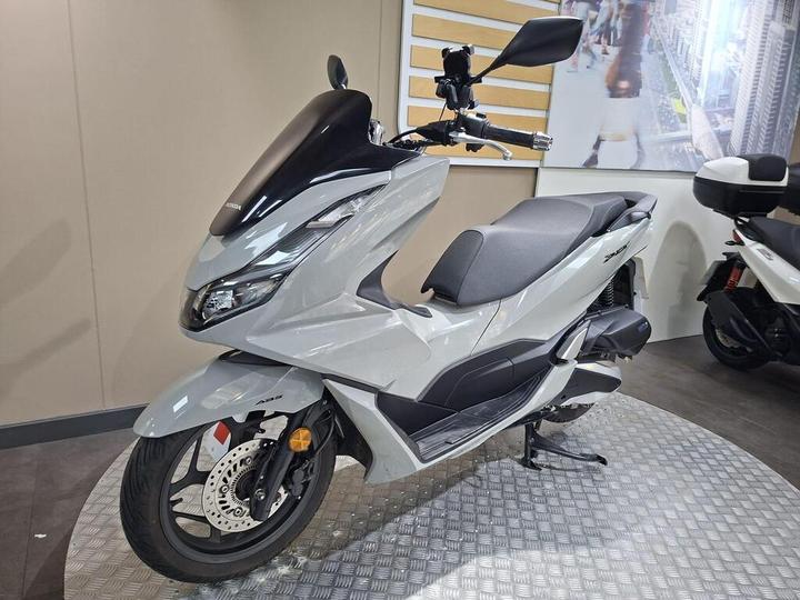 A sleek gray scooter displayed on a circular metallic platform, featuring a black windshield and a minimalist design, with a blurred background of an urban setting.