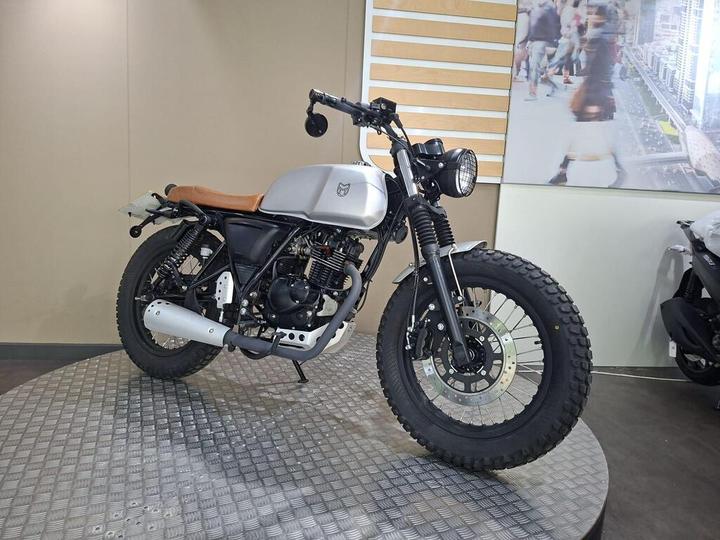 A custom motorcycle with a silver tank and brown leather seat, featuring rugged tires and a classic design, displayed on a textured platform against a neutral background.