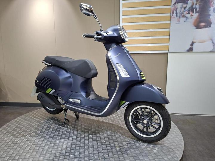 A blue Vespa scooter displayed in a showroom, featuring a sleek design with a black seat, alloy wheels, and subtle green stripes. The background includes neutral walls and a large image of a crowded street.