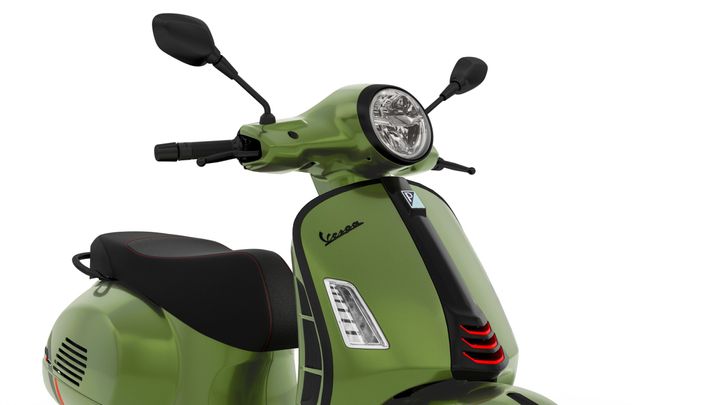 A close-up view of a green Vespa scooter highlighting its sleek design, featuring a round headlight, handlebars, and a stylish seat with red stitching.