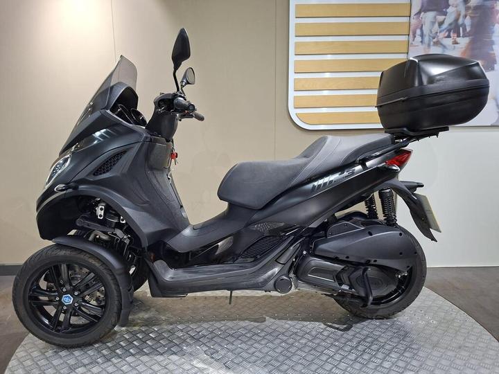 A black three-wheeled scooter with a windshield and rear storage box, displayed on a textured platform against a neutral wall.