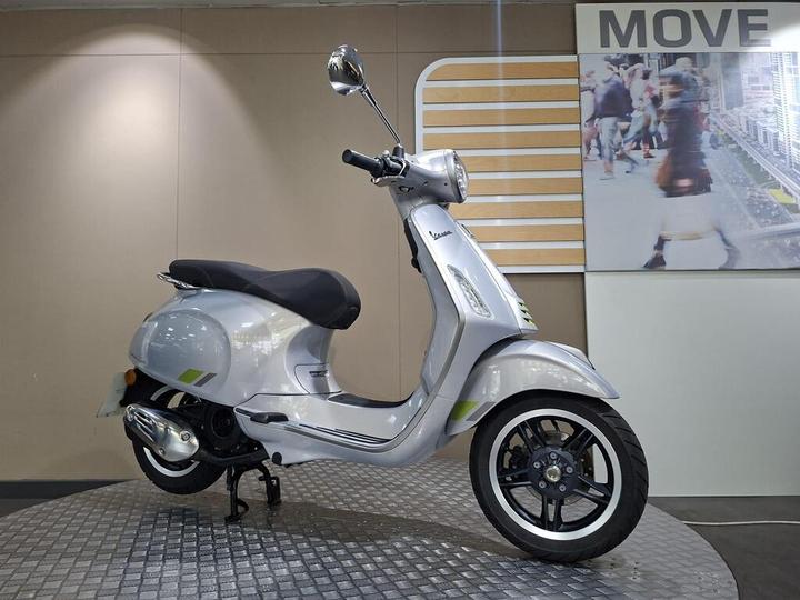 A silver Vespa scooter is showcased at an angle on a circular display platform, with a modern, minimalistic background featuring the word "MOVE" and an image of a bustling city scene.