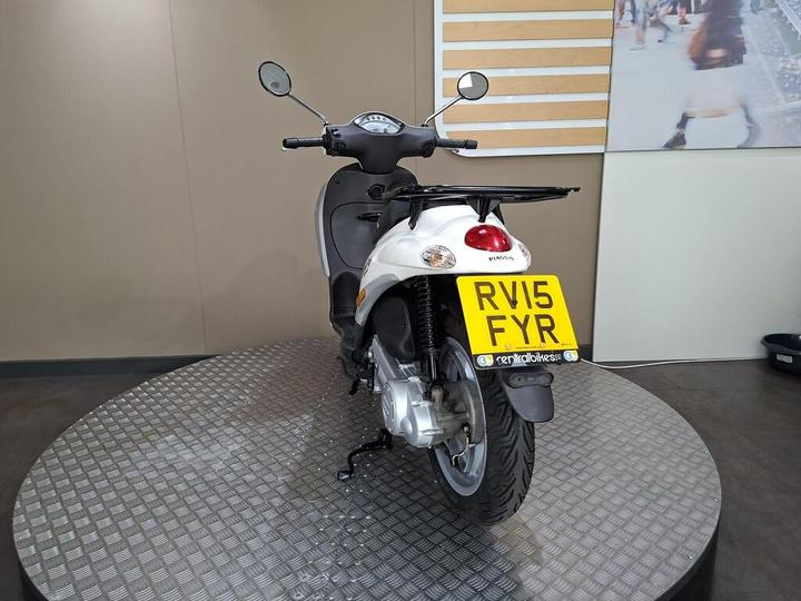 A white scooter displayed on a circular platform with a textured surface, featuring a rear view showing the license plate "RV15 FYR." A blurred image of pedestrians is visible in the background.