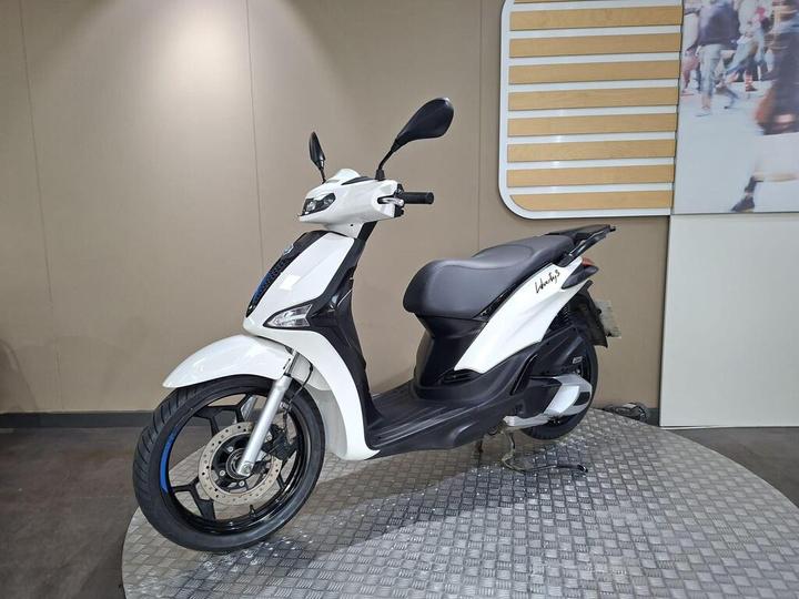 A white scooter displayed on a textured surface, against a neutral background with a wooden accent.