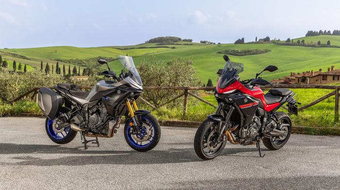 Two motorcycles are parked on a scenic road, surrounded by lush green hills and farmland. The motorcycle on the left is a silver model with blue wheels, and the one on the right is red with black accents. A rustic wooden fence and distant buildings are visible in the background under a clear sky.