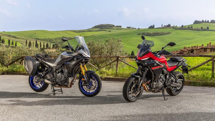 Two motorcycles are parked on a scenic road, surrounded by lush green hills and farmland. The motorcycle on the left is a silver model with blue wheels, and the one on the right is red with black accents. A rustic wooden fence and distant buildings are visible in the background under a clear sky.