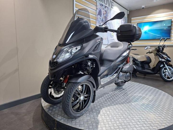 A black three-wheeled scooter displayed on a raised platform in a showroom, with a large windshield, storage box on the rear, and other scooters visible in the background.