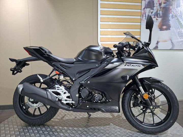 A black motorcycle with a sleek design is displayed on a textured surface, showcasing its sporty features, including a prominent exhaust, modern body shape, and detailed wheels, against a neutral background.
