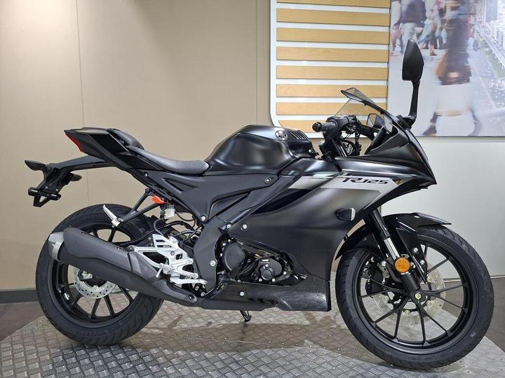 A black motorcycle with a sleek design is displayed on a textured surface, showcasing its sporty features, including a prominent exhaust, modern body shape, and detailed wheels, against a neutral background.