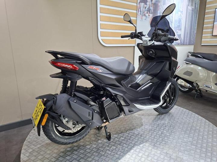A black scooter displayed on a rotating platform, showcasing its sleek design and modern features, with a blurred background of a showroom highlighting other vehicles.