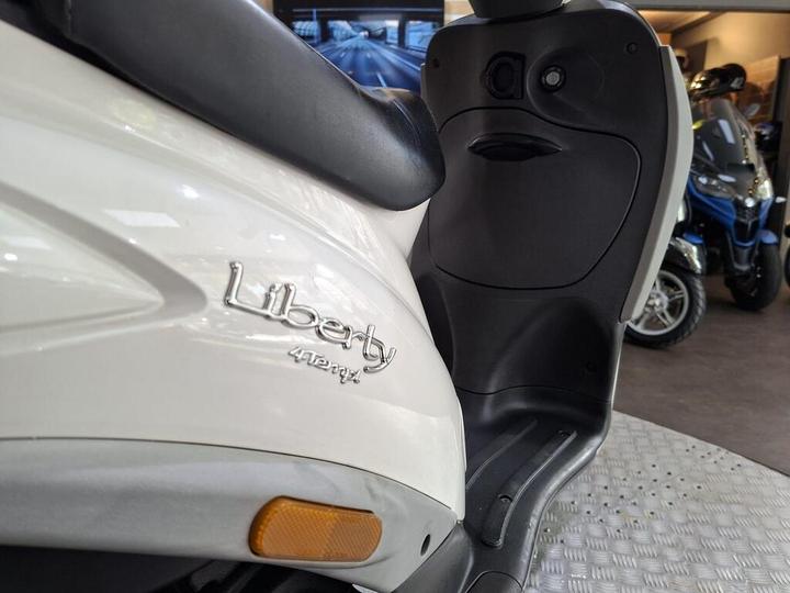 Close-up of a white scooter with the word "Liberty" and "125" emblem visible on the side, set against a backdrop of vehicles in a dealership showroom.