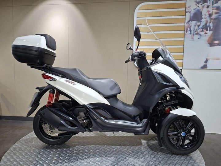 Piaggio MP3 300 HPE Sport (2022) A white and black three-wheeled scooter is parked on a circular metal platform in a showroom, featuring a top case and stand, with a neutral-colored wall and an abstract image of pedestrians in the background.