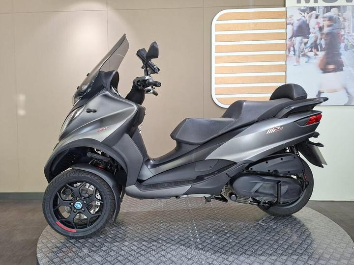 A sleek gray scooter with three wheels, featuring a tinted windshield and sporty black alloy wheels, displayed on a circular platform against a neutral background.
