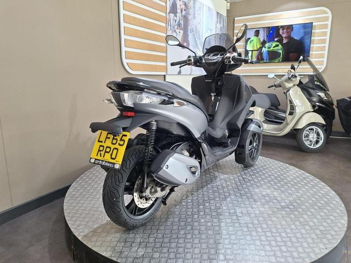 A gray scooter displayed on a circular platform in a showroom, with a license plate reading LF65 RPO, and a blurred background featuring a monitor and another scooter.