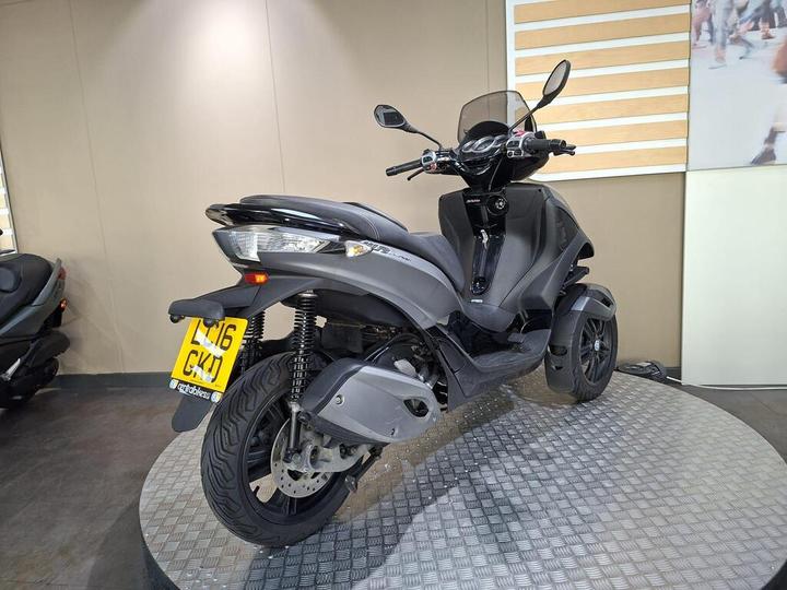 A gray three-wheeled scooter displayed on a circular platform in a showroom, featuring a modern design, front windshield, and visible license plate.