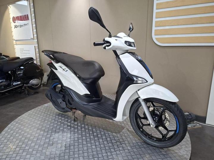 A white scooter with a sleek design, prominently featuring a blue accent, is displayed on a checkered platform in a showroom. The background shows a Yamaha logo and partially visible motorcycles.