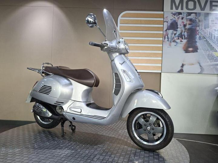A silver Vespa scooter with a brown seat, showcased on a textured surface in front of a promotional backdrop featuring blurred pedestrians and the word "MOVE."