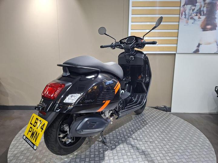 A black scooter with orange accents is displayed on a textured metallic surface in a showroom, showcasing its sleek design and features.