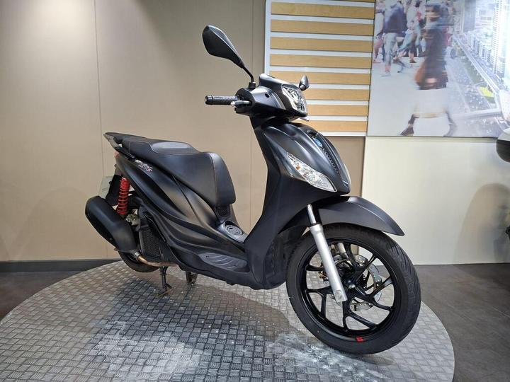 A sleek black scooter displayed on a circular metallic platform in a minimalist showroom, featuring a prominent front headlight and detailed wheel design. The background includes a subtle design element, enhancing the modern aesthetic.