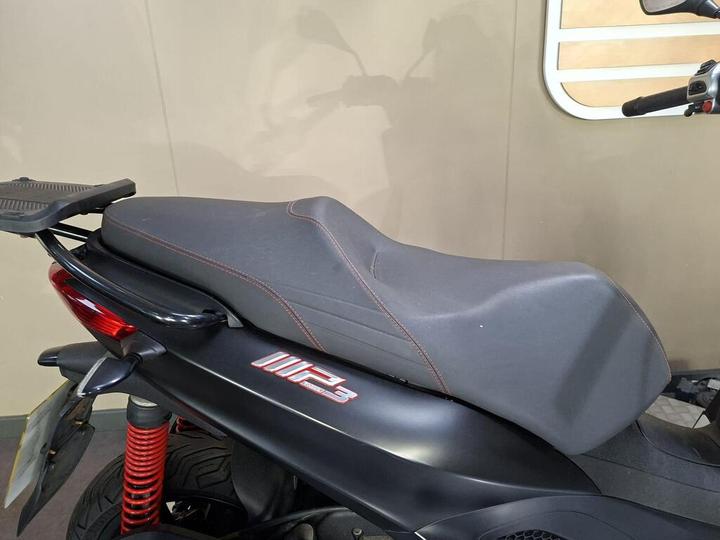 A close-up view of a motorcycle seat in black with red stitching, mounted on a motorcycle with a red rear shock absorber and visible parts of the bike's body.
