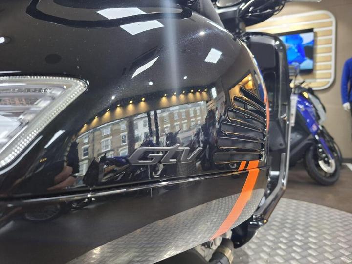 Close-up of a black motorcycle with a shiny surface reflecting lights and surroundings, featuring a prominent "GLV" emblem and orange accents.