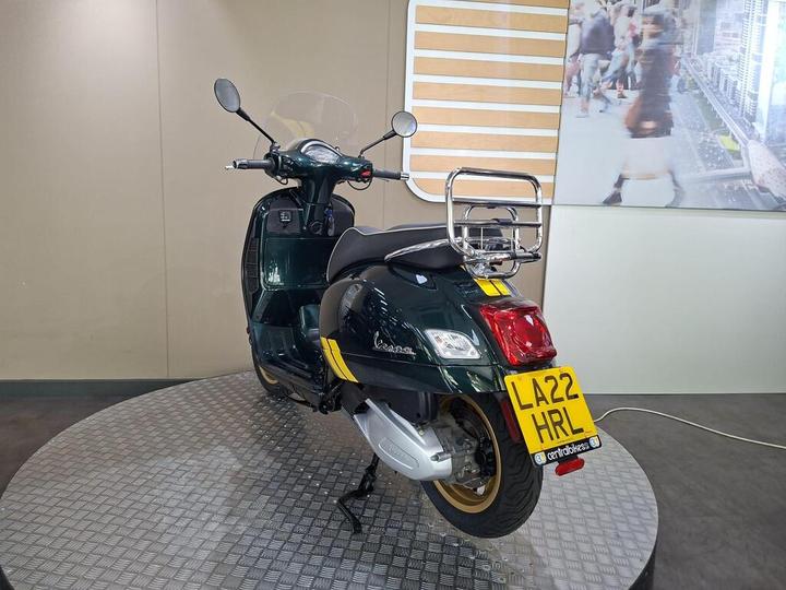 A green Vespa scooter displayed on a circular platform, viewed from the rear, featuring a chrome luggage rack and a visible license plate reading "LA22 HRL." The background includes a neutral-colored wall.