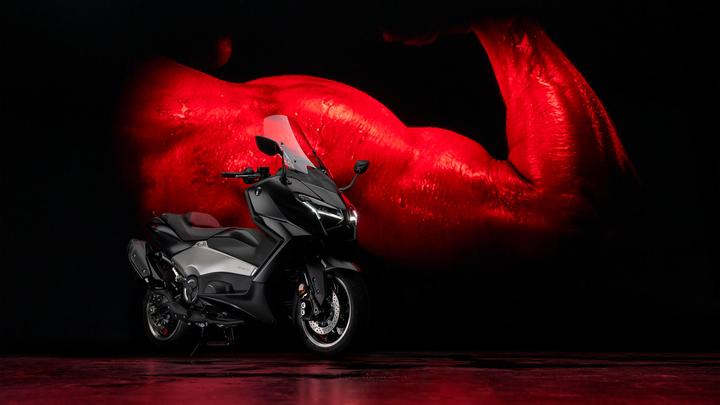 A sleek black and silver motorcycle is positioned against a dark background featuring a red-hued muscular arm. The bike is highlighted with dramatic lighting, emphasizing its modern design.