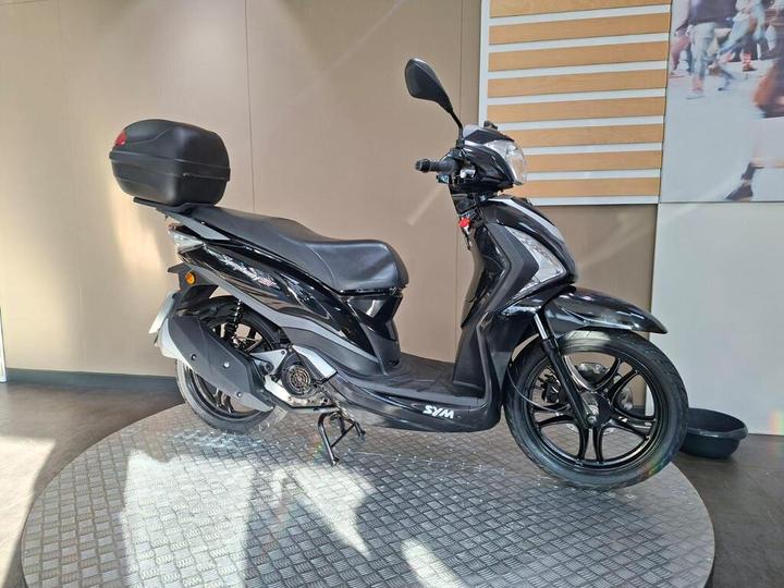 A black SYM scooter on a circular display stand in a well-lit showroom, with a rear storage compartment and a sleek design.